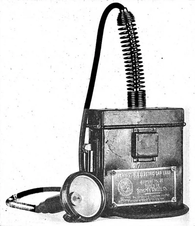 Black-and-white photo of a 1950 electric miners’ cap lamp with a separate rechargeable battery pack made by Concordia, Cardiff. The lamp is helmet-mounted and connected by a cable, reducing flame risks in mines.