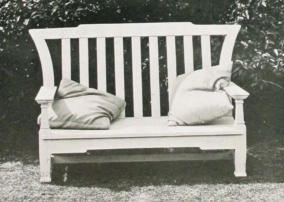 Historic black-and-white photograph of a two-seat wooden garden bench with a tall vertical-slatted backrest, gently flared upper corners, square armrests, low seat, straight legs, and several loose cushions, standing on a lawn in front of dense foliage.