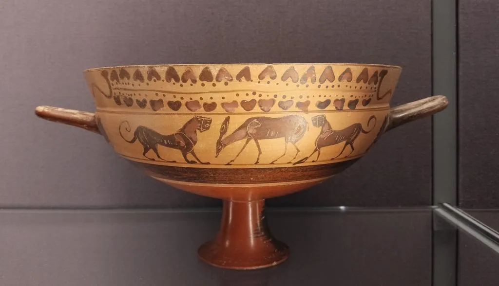 Ancient Corinthian kylix with painted animal hunt scene, featuring lions attacking a grazing deer, surrounded by decorative patterns.