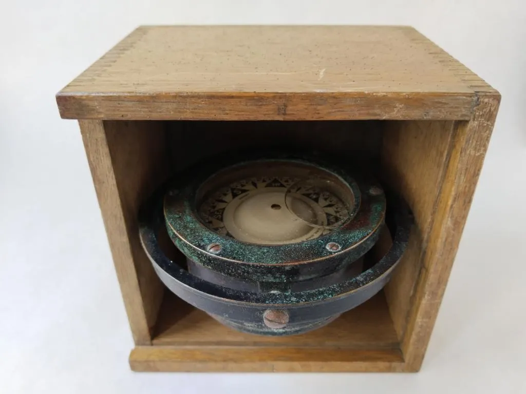 Antiker Nasskompass in einer hölzernen Schutzhülle, mit sichtbaren Spuren von Alter und Gebrauch. Der Kompass zeigt eine deutliche grüne Patina und verwendet ein gyroskopisches System zur Stabilisierung der Nadel.