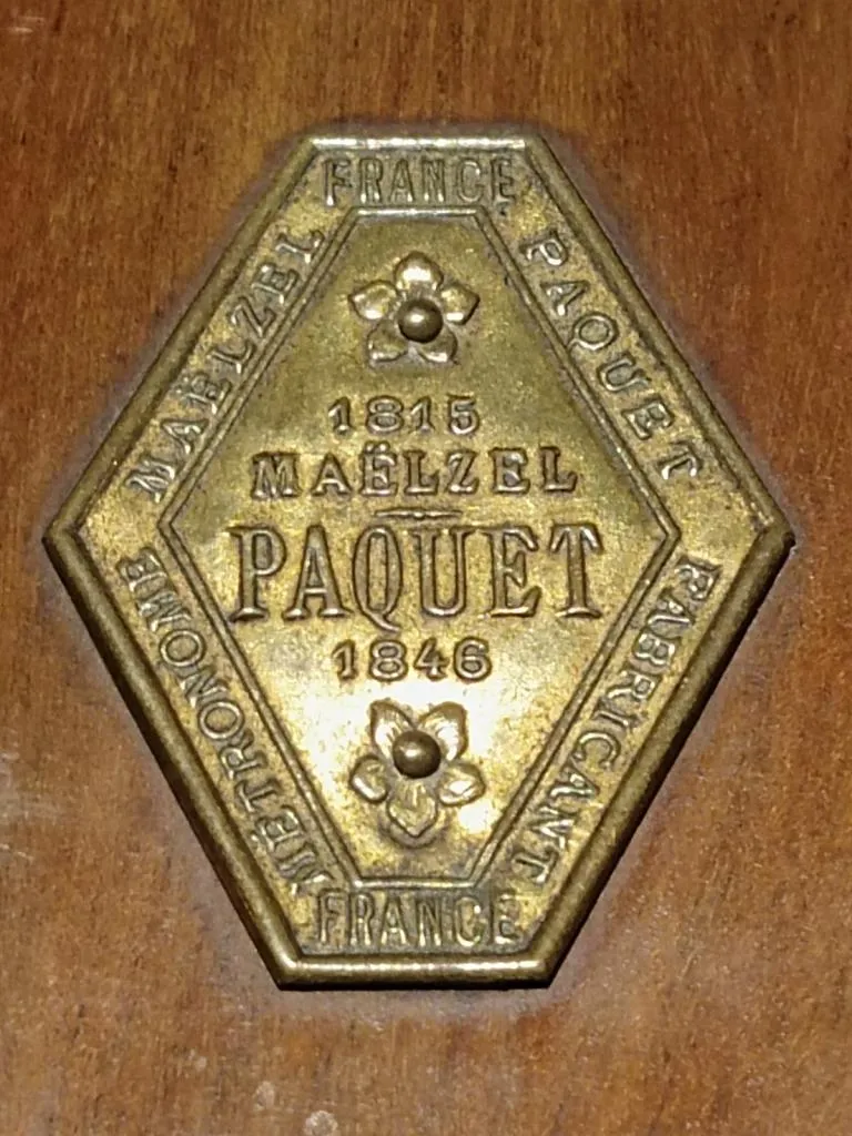 Brass maker's plaque from a Maelzel Metronome. The hexagonal plate is inscribed with 1815 Maelzel PAQUET 1846, along with the words Metronome, France, and Fabricant, indicating its origin and manufacturer.