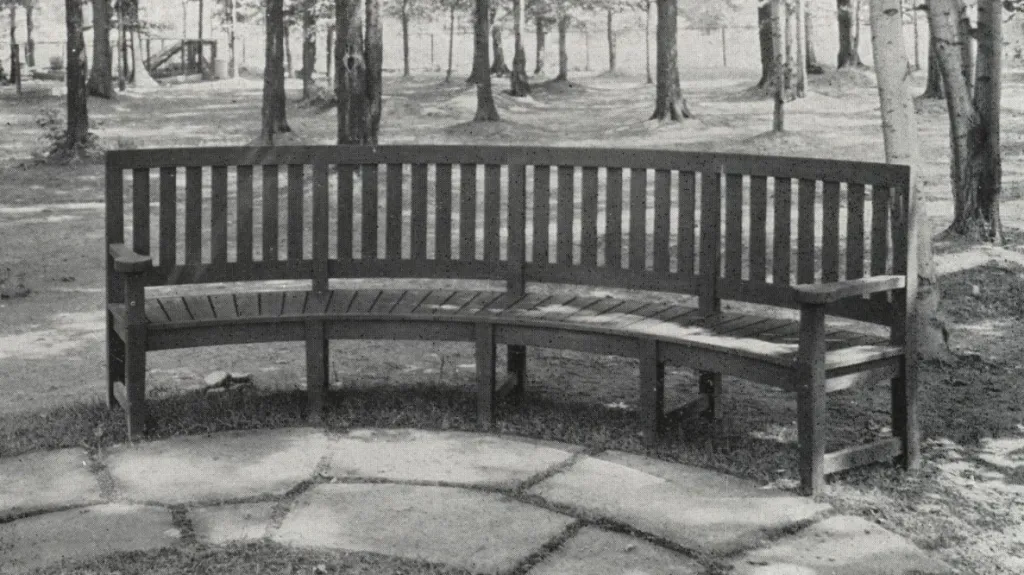 Black-and-white catalog photograph of a large curved wooden garden bench with a gently arched slatted backrest, curved slatted seat, wide armrests, and multiple support legs, positioned on a stone-paved area beneath mature trees in a landscaped garden.