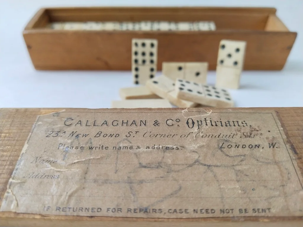 Close-up of an antique wooden domino box with a worn paper label reading Callaghan & Co. Opticians, 23a New Bond St., corner of Conduit St., London, W. and bone domino tiles blurred in the background.