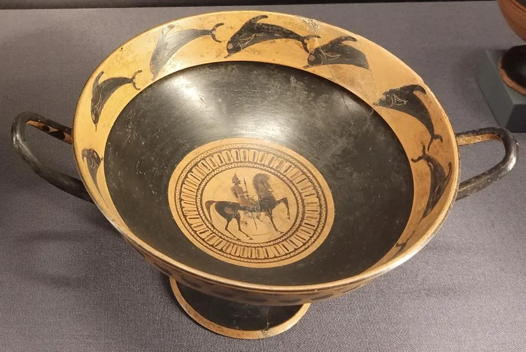 Ancient East Greek kylix with two handles. Inside: central tondo showing a mounted warrior, rim decorated with leaping dolphins. Ca. 550 BCE.