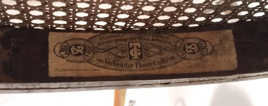 Nahaufnahme eines frühen Thonet-Papieretiketts an der Unterseite eines Stuhls mit Rohrgeflecht, Beschriftung Gebrüder Thonet Wien mit verzierten Buchstaben und dekorativem Rahmen..