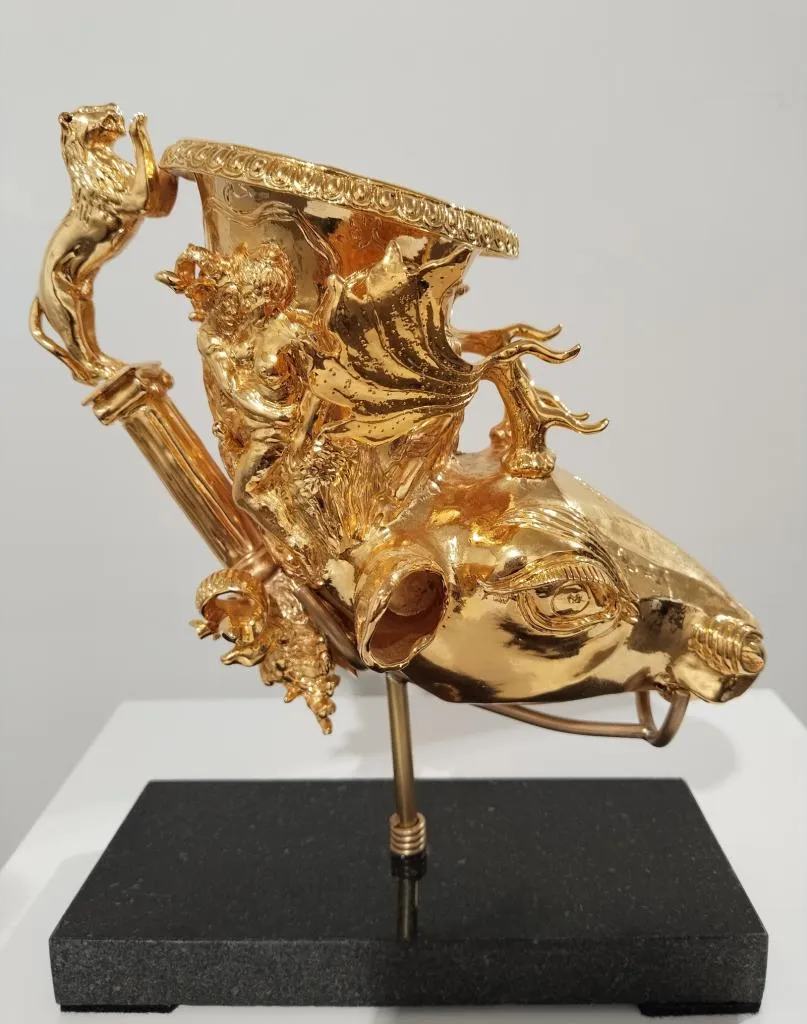 Zeremonielles Trinkgefäß aus Gold in Form eines Widderkopfes, mit kunstvollen mythologischen Reliefs und einem löwenförmigen Griff, auf schwarzem Sockel präsentiert.
