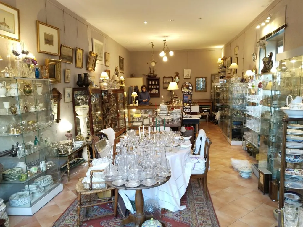Inside L’Estive Antiques: crystal glassware, fine porcelain, and vintage treasures beautifully arranged — with Madame Isabelle Ripert-Grac welcoming guests from the counter.