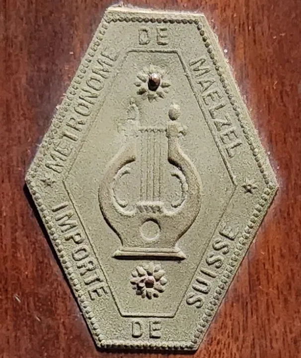 Close-up detail of a hexagonal metal plaque mounted on the wooden case of an antique Swiss Paillard & Cie Maelzel metronome, showing a lyre motif and the inscriptions Métronome de Maelzel and Importé de Suisse.