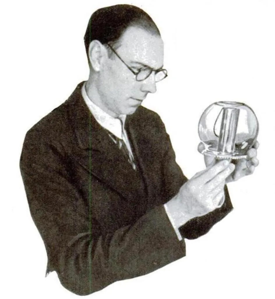 Ein Mann im Anzug und mit Brille hält ein Modell der Davy-Sicherheitslampe in eine Glasschale, um deren Sicherheit in brennbaren Gasatmosphären zu demonstrieren.