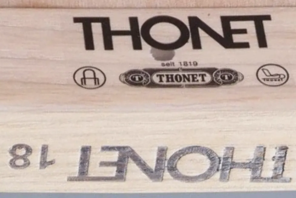 Close-up of a modern Thonet marking on a wooden frame, showing a transparent sticker with bold THONET lettering, old emblem design, date 'seit 1819', and symbols, together with an inked stamp of the logo and model number.