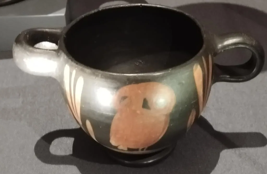 Black-glazed ancient Greek skyphos with a red-figure depiction of an owl, flanked by vertical decoration; two handles on each side.