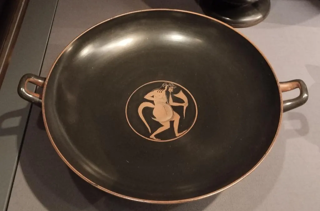 Attic red-figure kylix showing a mythological Silen with a drinking horn in the center, black glazed with two handles.