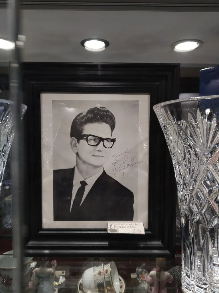 This is a signed photo of Roy Orbison. The black and white photograph is framed in a black frame and features Roy Orbison wearing his iconic glasses and a suit. His signature, Sincerely, Roy Orbison, is visible on the right side of the photo. The photograph is displayed in a glass cabinet alongside various other items, including crystal glassware and porcelain, highlighting its placement in an antique setting.