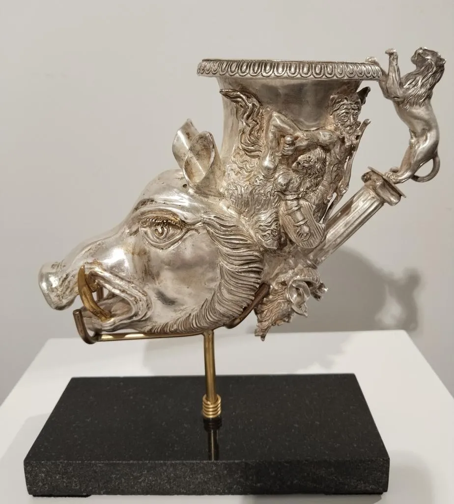 Zeremonielles Trinkgefäß aus Silber in Form eines Eberkopfes mit Reliefs von Löwen und mythologischen Figuren, auf einem schwarzen Sockel präsentiert.