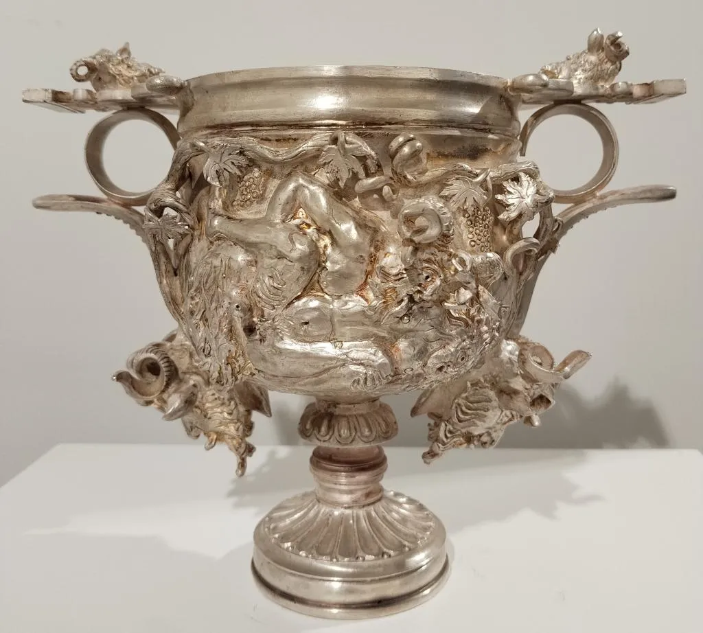 Decorative silver skyphos with high-relief figures, grape clusters, and mythological motifs in Dionysian style; two loop handles and a fluted pedestal base.