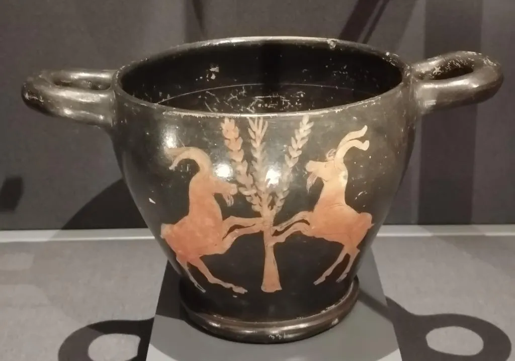 Antiker rotfiguriger Skyphos mit zwei aufgerichteten Ziegenböcken an einem zentralen Baummotiv; schwarz glasiert mit zwei Henkeln.