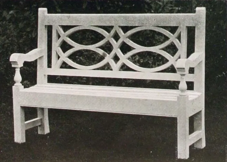 Black-and-white photograph of a small wooden garden bench with a straight slatted seat, square legs, shaped armrests with turned supports, and an ornamental open back formed by symmetrical interlacing curved wooden elements.