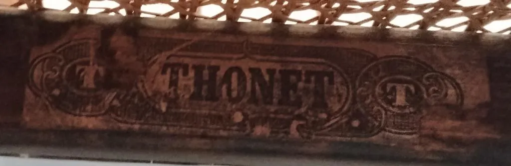 Nahaufnahme eines späteren Thonet-Papieretiketts unter einer Sitzfläche aus Rohrgeflecht, mit der Aufschrift THONET in kräftiger Schrift und ornamentalen T-Symbolen auf beiden Seiten.