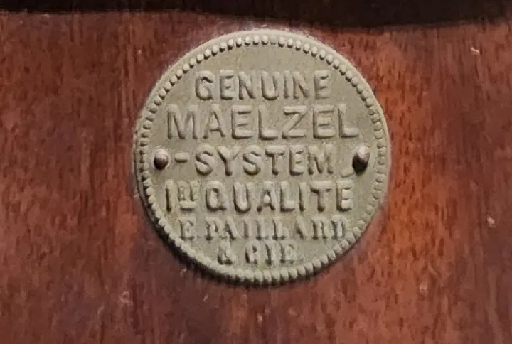 Close-up detail of a round metal label mounted on the wooden case of an antique Swiss Paillard & Cie Maelzel metronome, reading “Genuine Maelzel System, 1re Qualité, and E. Paillard & Cie.