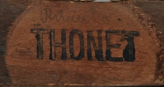 Nahaufnahme eines flachen schwarzen Tintenstempels mit der Aufschrift THONET auf einem Sitzrahmen aus Holz, mit Abnutzung und ungleichmäßigem Farbauftrag.
