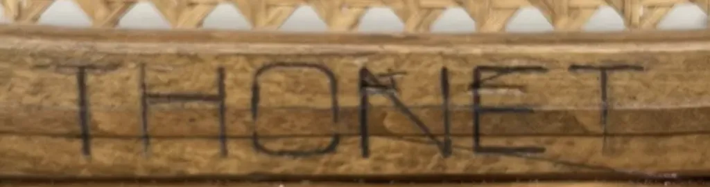 Close-up of a flat ink stamp reading THONET in slim, modern-style lettering on a wooden chair frame beneath a cane seat.
