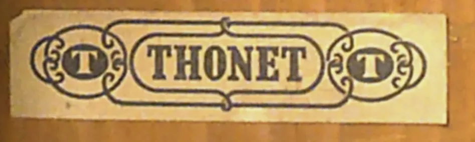 Nahaufnahme eines vereinfachten Thonet-Papieretiketts auf Holz, mit der schwarzen Aufschrift THONET in kräftigen Buchstaben, flankiert von dekorativen T-Symbolen.