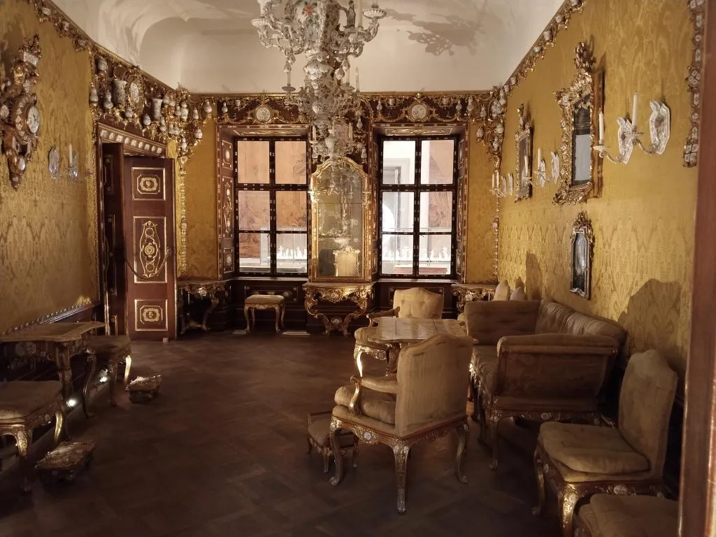 Ein königlicher Saal mit antiken Sesseln, einem alten Holztisch mit Marmorplatte. Die Szene umfasst ein antikes Möbelstück und einen antiken Spiegel.