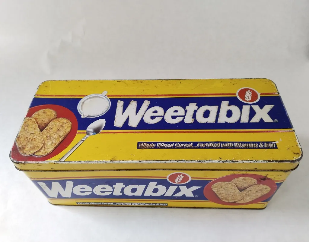 An old, well-worn tin box with a bright yellow and blue design, featuring a spoon and wheat biscuits on a red plate, hinting at its past use as a storage container.