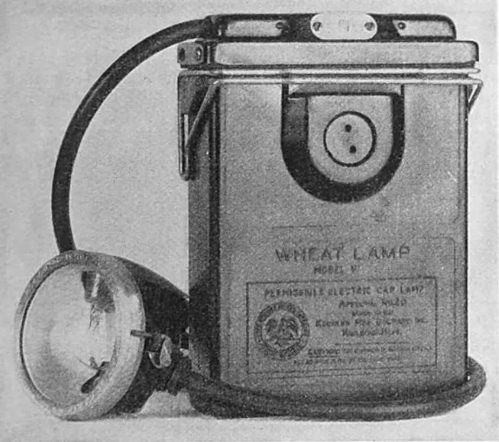 Black-and-white photograph of a Wheat electric cap lamp with a belt-mounted battery pack and wired headlamp, manufactured by Koehler Manufacturing Company in the United States.