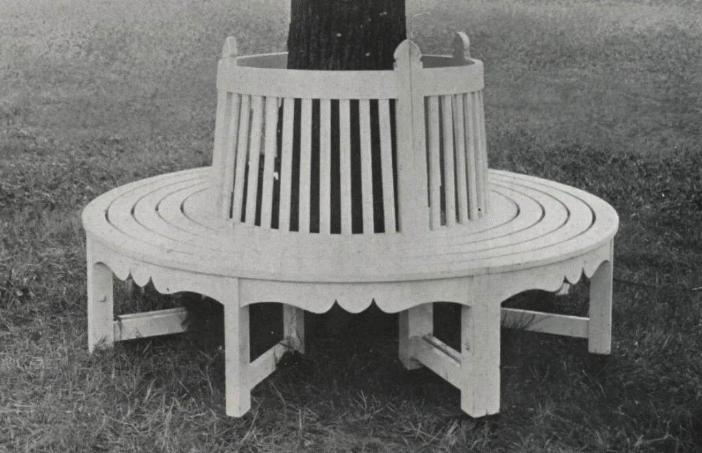 Black-and-white catalog photograph of a circular wooden tree bench surrounding a mature tree, featuring a continuous slatted seat, vertical slatted inner backrest, and decorative scalloped apron in a classic park and garden design.