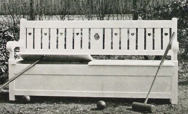 Black-and-white photograph of a wooden garden bench with a straight slatted backrest, small decorative cutouts, broad armrests, and a large enclosed storage compartment beneath the hinged seat, with croquet mallets and balls arranged around the bench.