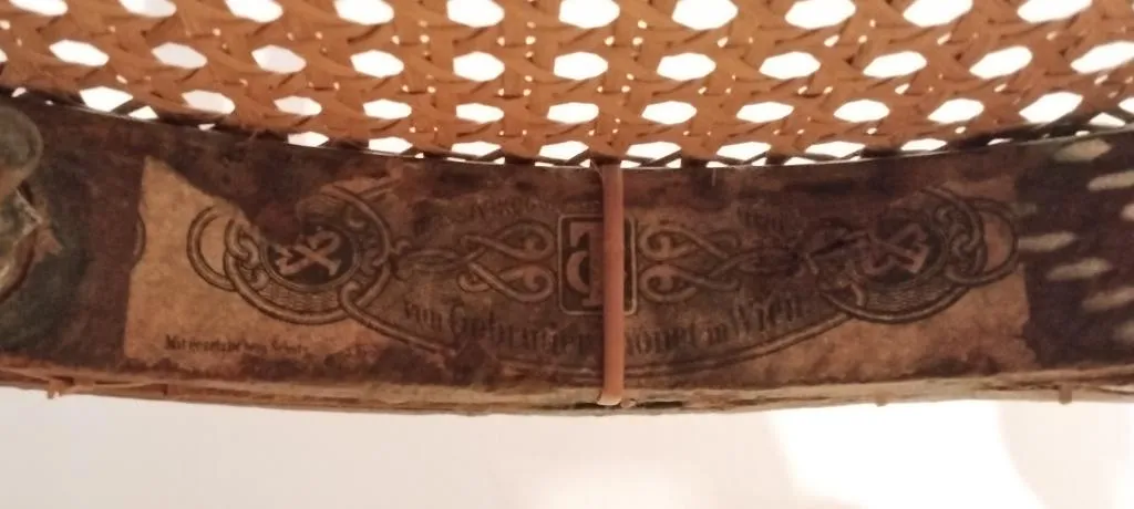 Close-up of a faded Thonet paper label under a cane seat, showing decorative border and partially visible Gebrüder Thonet Wien text.
